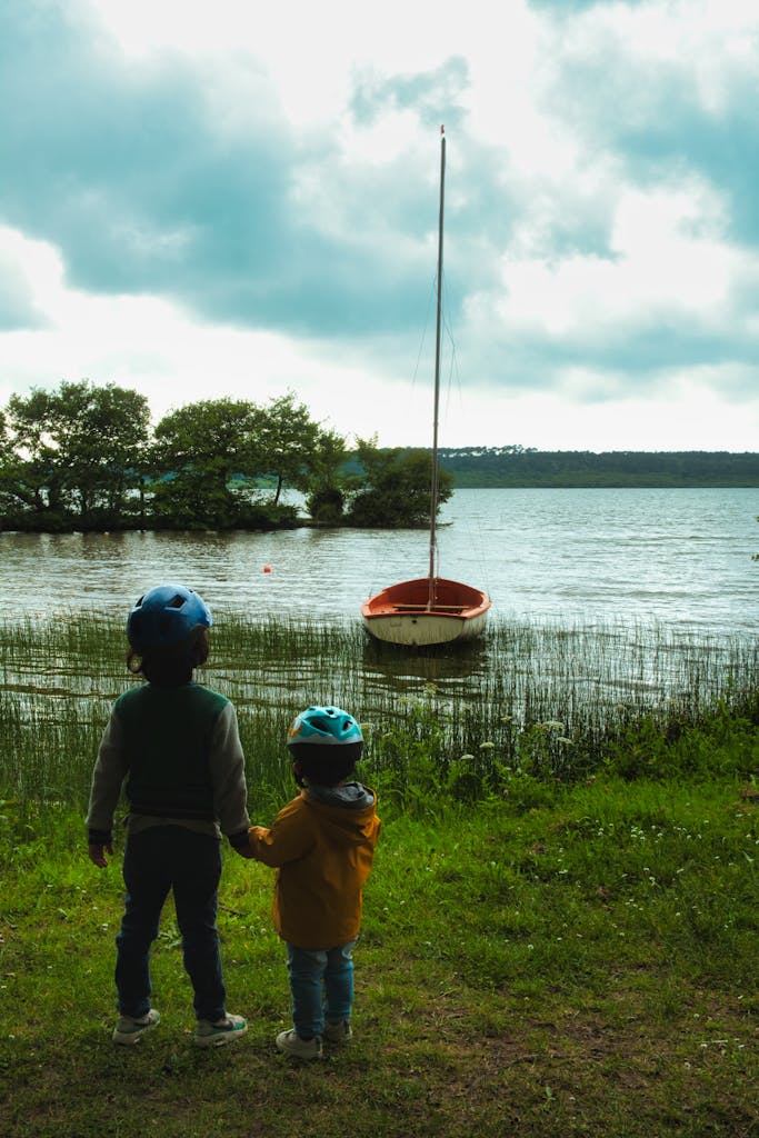 Two children wearing helmets, holding hands, gaze at a lake sailboat.